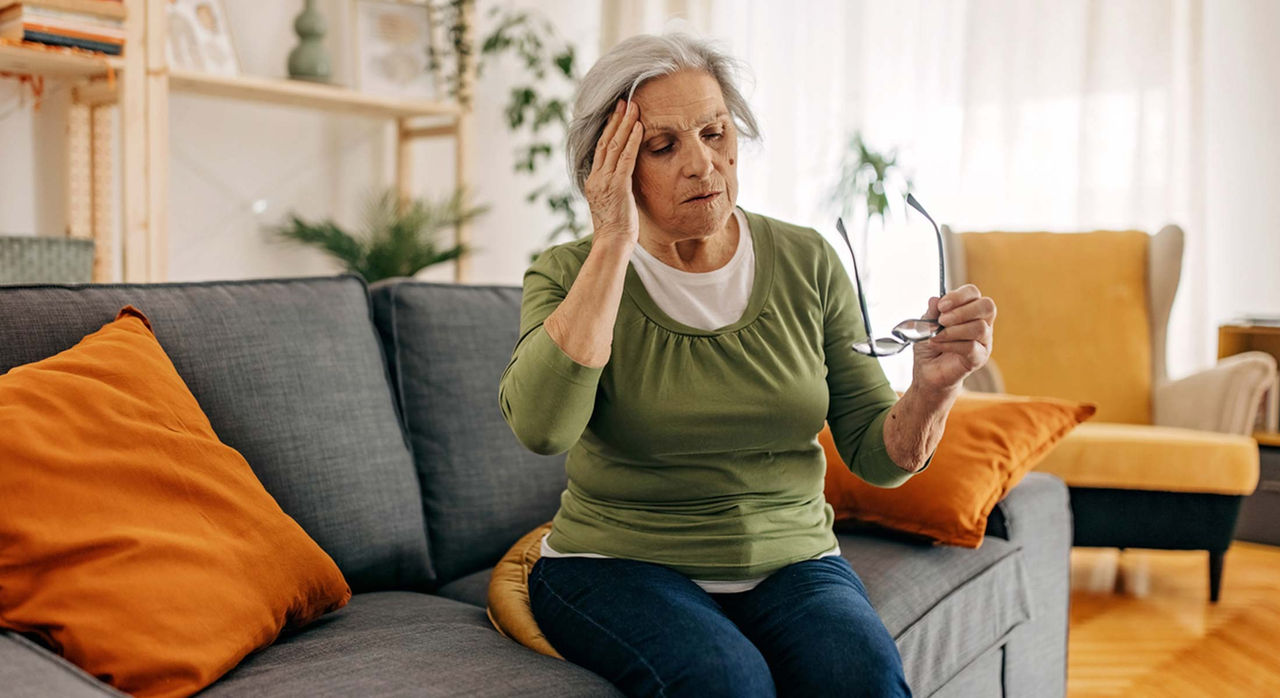 An older woman with white hair puts up her right hand on the right side of her forehead while holding her glasses in her left hand. She is wearing white t-shirt with an earthy green long-sleeve t-shirt over it, and blue denim jeans. She is sitting on a blue couch in a living room.